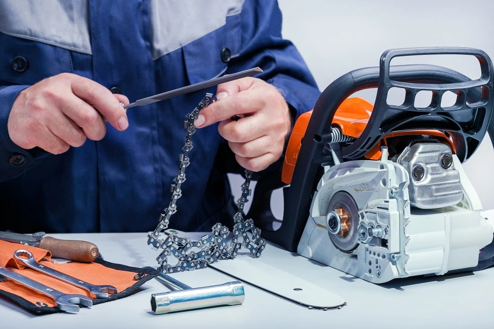 Person in blue jacket sharpening a chainsaw chain with a file, tools laid out